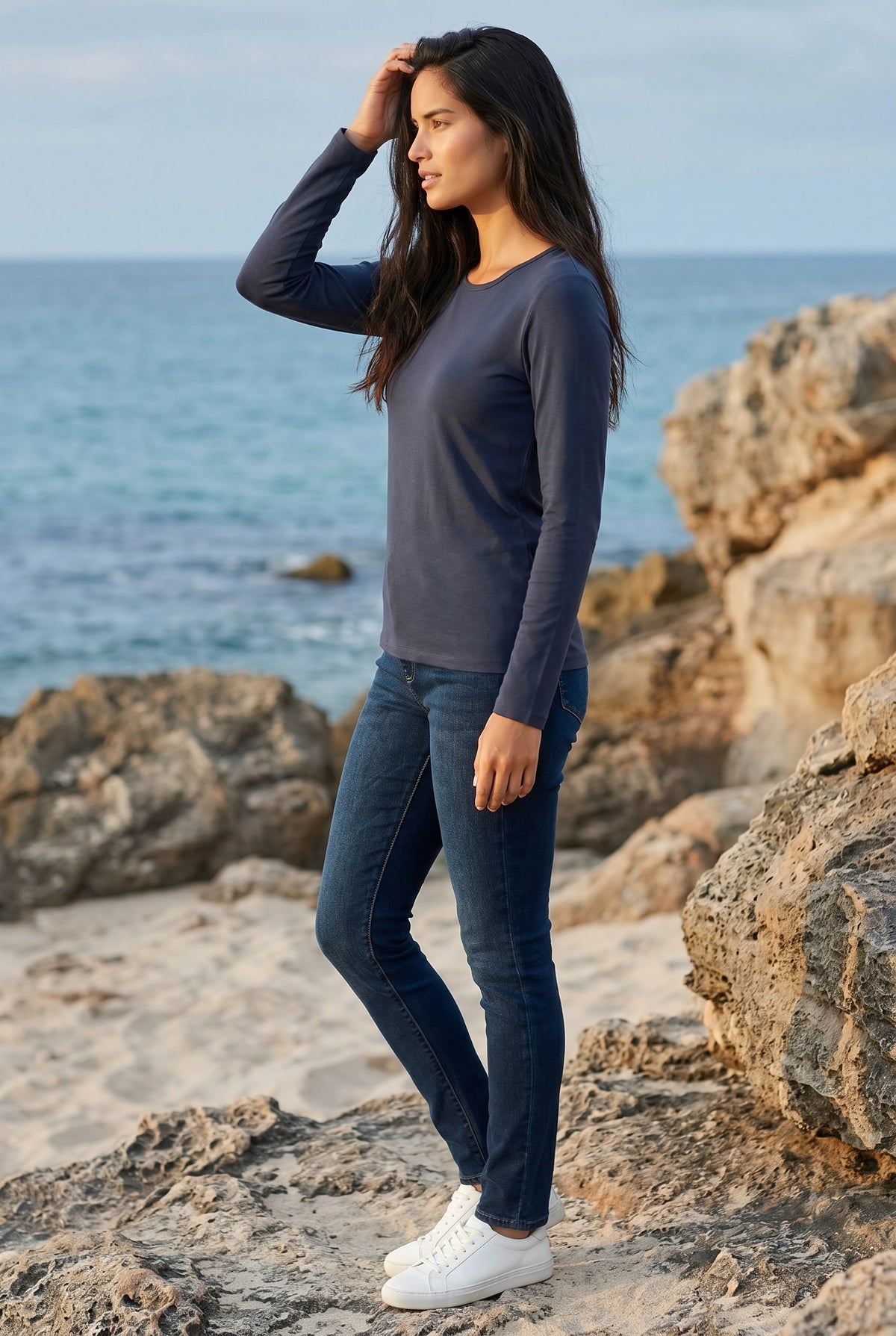 Woman in steel blue long sleeve Pima cotton tee and jeans standing on a rocky beach